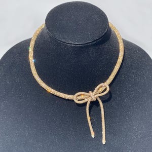 May include: A gold-toned choker necklace featuring a flexible, tubular mesh band covered in sparkling crystals. The necklace includes a prominent, integrated bow-shaped pendant at the centre, with two dangling ends that extend downward from the knot.