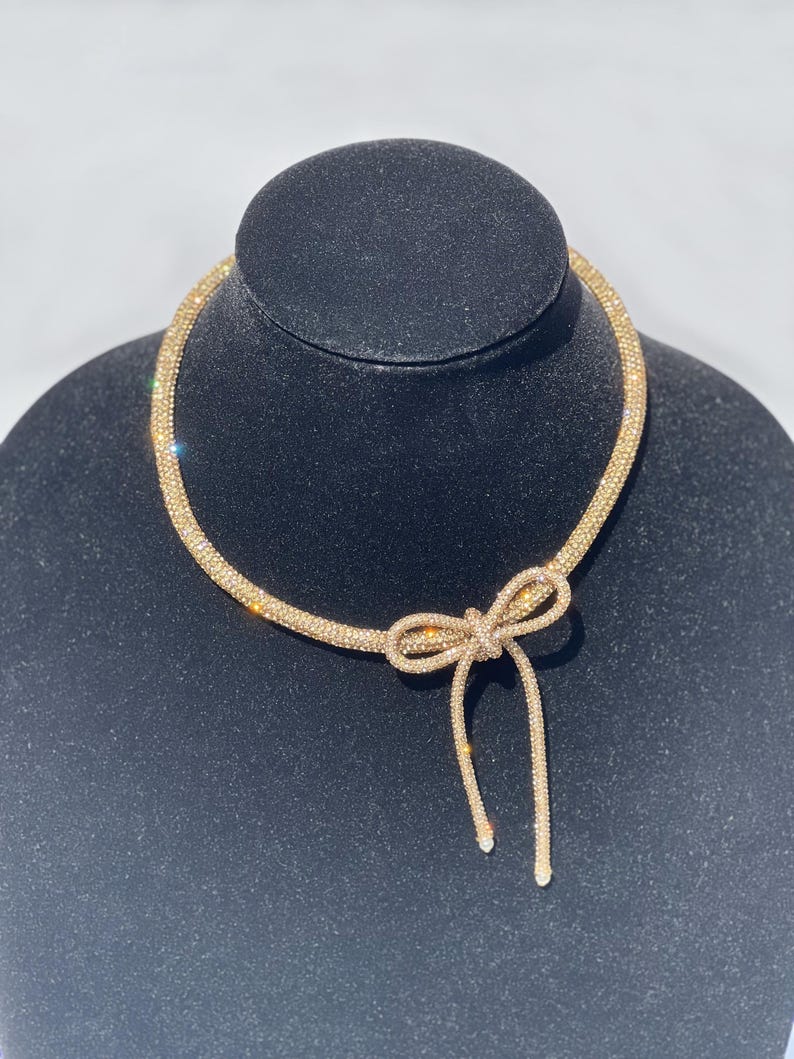 May include: A gold-toned choker necklace featuring a flexible, tubular mesh band covered in sparkling crystals. The necklace includes a prominent, integrated bow-shaped pendant at the centre, with two dangling ends that extend downward from the knot.