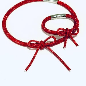 May include: Two red glittery cord bracelets featuring a sparkling texture and a decorative bow tied at the front. Each bracelet is secured with a polished silver-toned cylindrical metal clasp, creating a cohesive set of matching accessories.