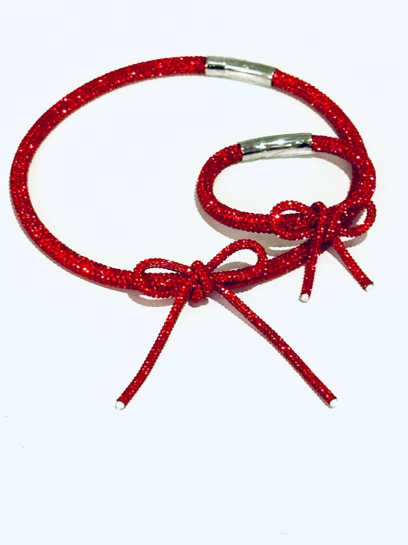May include: Two red glittery cord bracelets featuring a sparkling texture and a decorative bow tied at the front. Each bracelet is secured with a polished silver-toned cylindrical metal clasp, creating a cohesive set of matching accessories.