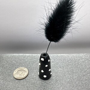 Puede incluir: Un peque&ntilde;o jarr&oacute;n de cer&aacute;mica negro y blanco con lunares con una sola pluma negra que sobresale de la parte superior.