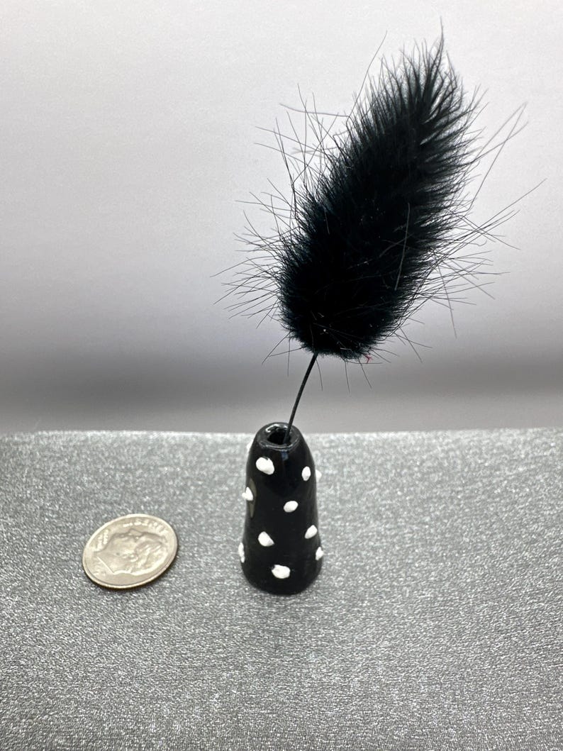Puede incluir: Un peque&ntilde;o jarr&oacute;n de cer&aacute;mica negro y blanco con lunares con una sola pluma negra que sobresale de la parte superior.