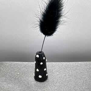 Puede incluir: Un peque&ntilde;o jarr&oacute;n de cer&aacute;mica negro con lunares blancos contiene una sola pluma negra.