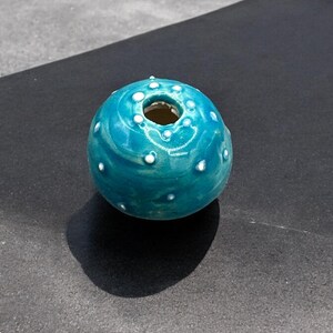 Op de afbeelding: Een kleine, tealblauwe keramische vaas met witte stippen en een swirl-patroon. De vaas heeft een ronde vorm en een kleine opening aan de bovenkant.