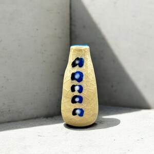 May include: A small, cream-colored ceramic vase with a crackled glaze. The vase has a blue and black design with a series of circles.