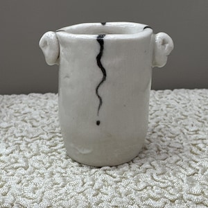 May include: A small, off-white ceramic vessel with a cylindrical body and two small, rounded handles on the sides. The surface features a single, thin, vertical black wavy line that extends from the top rim down to a small dot near the base.