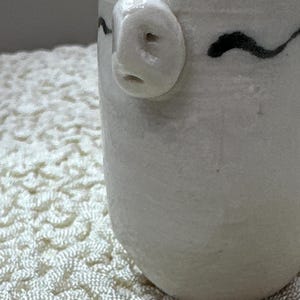 May include: A small, off-white ceramic vessel with a cylindrical shape and a smooth, glossy finish. The rim features hand-painted black wavy lines, and a small, sculpted, three-dimensional face with a nose and mouth is attached to the upper side.