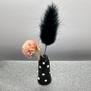 Puede incluir: Un peque&ntilde;o jarr&oacute;n de cer&aacute;mica negro con lunares blancos contiene una flor rosa seca y una pluma negra.