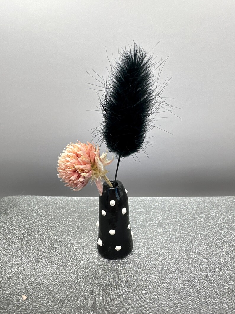 Puede incluir: Un peque&ntilde;o jarr&oacute;n de cer&aacute;mica negro con lunares blancos contiene una flor rosa seca y una pluma negra.