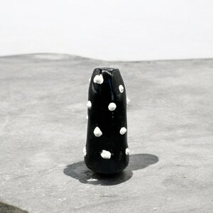 Könnte beinhalten: Eine schwarze Keramikvase mit weißen Punkten. Die Vase ist hoch und schlank, mit abgerundetem Ober- und Unterteil.