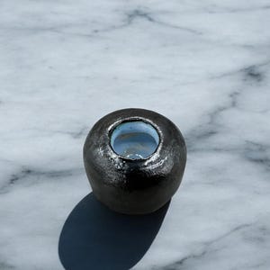 May include: A small, black ceramic vase with a round shape and a smooth, slightly textured surface. The vase has a small opening at the top.