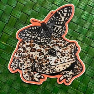 May include: A sticker featuring a cartoon illustration of a brown and white toad with a black and white butterfly on its head. The toad has a spotted pattern and the butterfly has black and white wings.