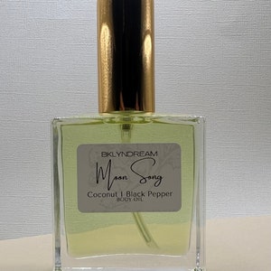 May include: A clear glass body oil bottle with a gold cap. The label reads "BKLYNDREAM Moon Song Coconut & Black Pepper Body Oil". The liquid inside is a pale yellow color. The bottle has a rectangular shape.