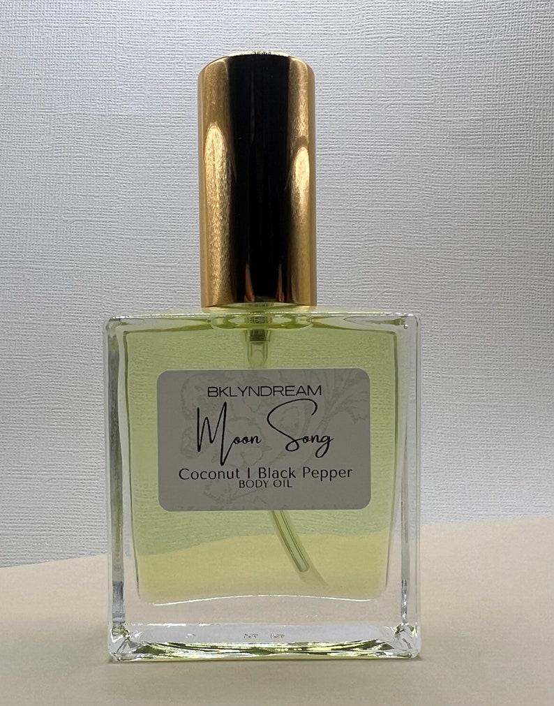 May include: A clear glass body oil bottle with a gold cap. The label reads "BKLYNDREAM Moon Song Coconut & Black Pepper Body Oil". The liquid inside is a pale yellow color. The bottle has a rectangular shape.