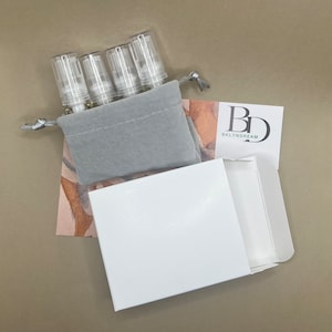 May include: Four small clear glass bottles with gold lids and a white label are in a gray velvet pouch with a drawstring. The pouch is on top of a white box with a white lid. The box is open and the lid is off to the side. A white card with the text "BD BKLYNDREAM" is in the background.