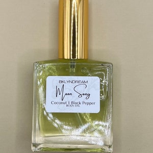 May include: A clear glass bottle of body oil with a gold cap. The oil inside is a light green color. A white label on the front reads "BKLYNDREAM Moon Song Coconut | Black Pepper BODY OIL."