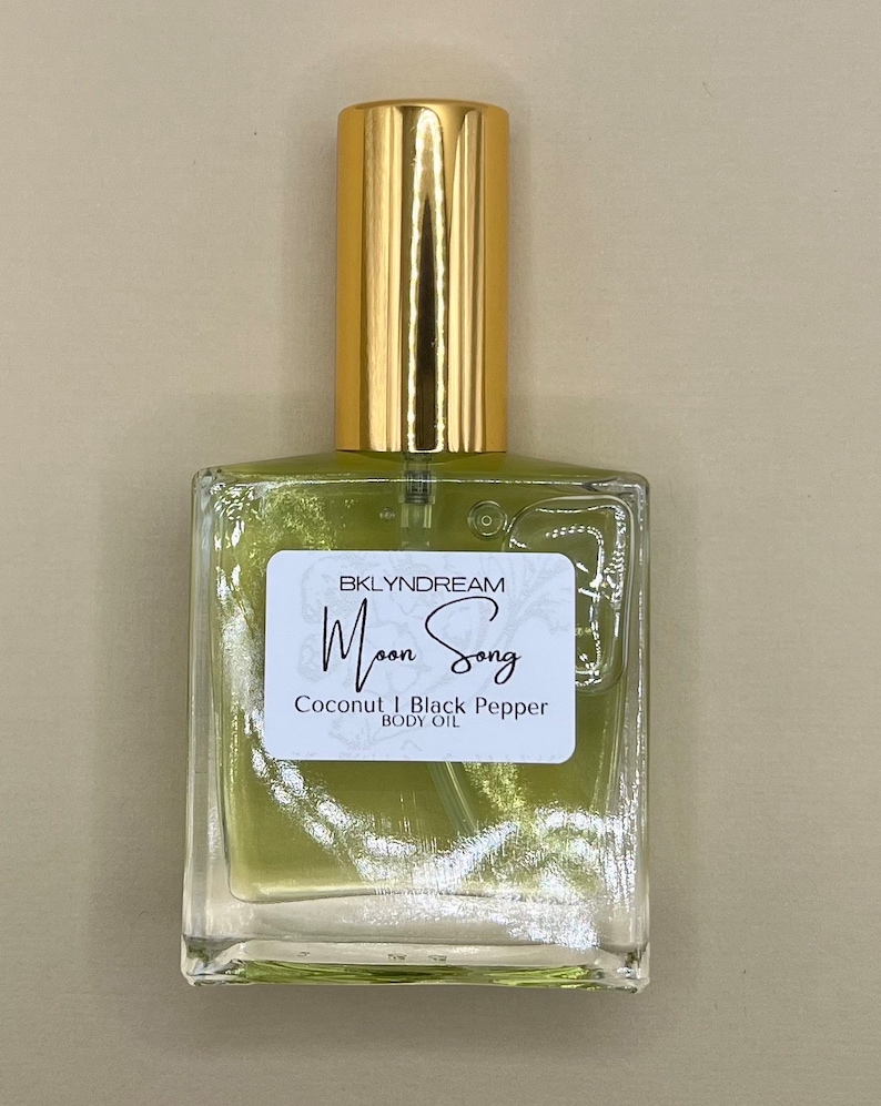 May include: A clear glass bottle of body oil with a gold cap. The oil inside is a light green color. A white label on the front reads "BKLYNDREAM Moon Song Coconut | Black Pepper BODY OIL."