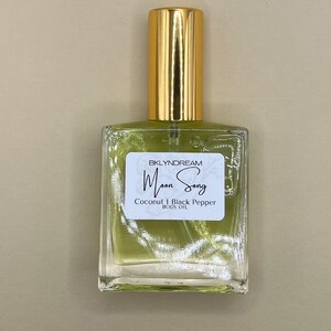 May include: A square glass bottle of body oil with a gold cap. The label reads "Moon Song" and lists "Coconut & Black Pepper" as ingredients. The oil inside is a light green color.