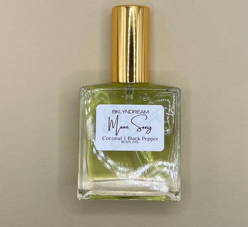 May include: A square glass bottle of body oil with a gold cap. The label reads "Moon Song" and lists "Coconut & Black Pepper" as ingredients. The oil inside is a light green color.