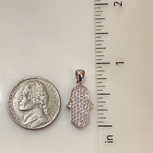 May include: A silver-colored Hamsa hand pendant, encrusted with small, clear gemstones. The pendant is approximately 1 inch in length. A U.S. quarter coin is next to the pendant for size comparison. The pendant is on a white surface.