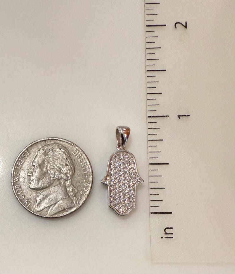 May include: A silver-colored Hamsa hand pendant, encrusted with small, clear gemstones. The pendant is approximately 1 inch in length. A U.S. quarter coin is next to the pendant for size comparison. The pendant is on a white surface.