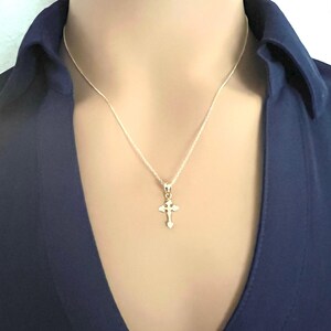 May include: A silver cross pendant necklace. The cross is small and detailed, suspended from a delicate silver chain. The person is wearing a dark blue collared shirt.