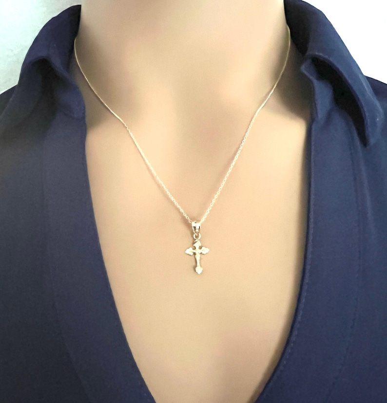 May include: A silver cross pendant necklace. The cross is small and detailed, suspended from a delicate silver chain. The person is wearing a dark blue collared shirt.