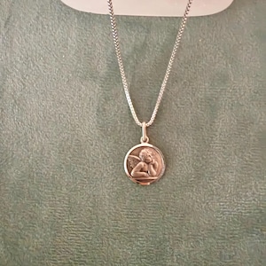 Peut inclure: Collier argent&eacute; avec un pendentif rond repr&eacute;sentant un ch&eacute;rubin en relief. Le ch&eacute;rubin a des ailes et repose sa t&ecirc;te sur sa main. La cha&icirc;ne est de conception fine et tiss&eacute;e. Le pendentif et la cha&icirc;ne sont pos&eacute;s sur un fond vert.