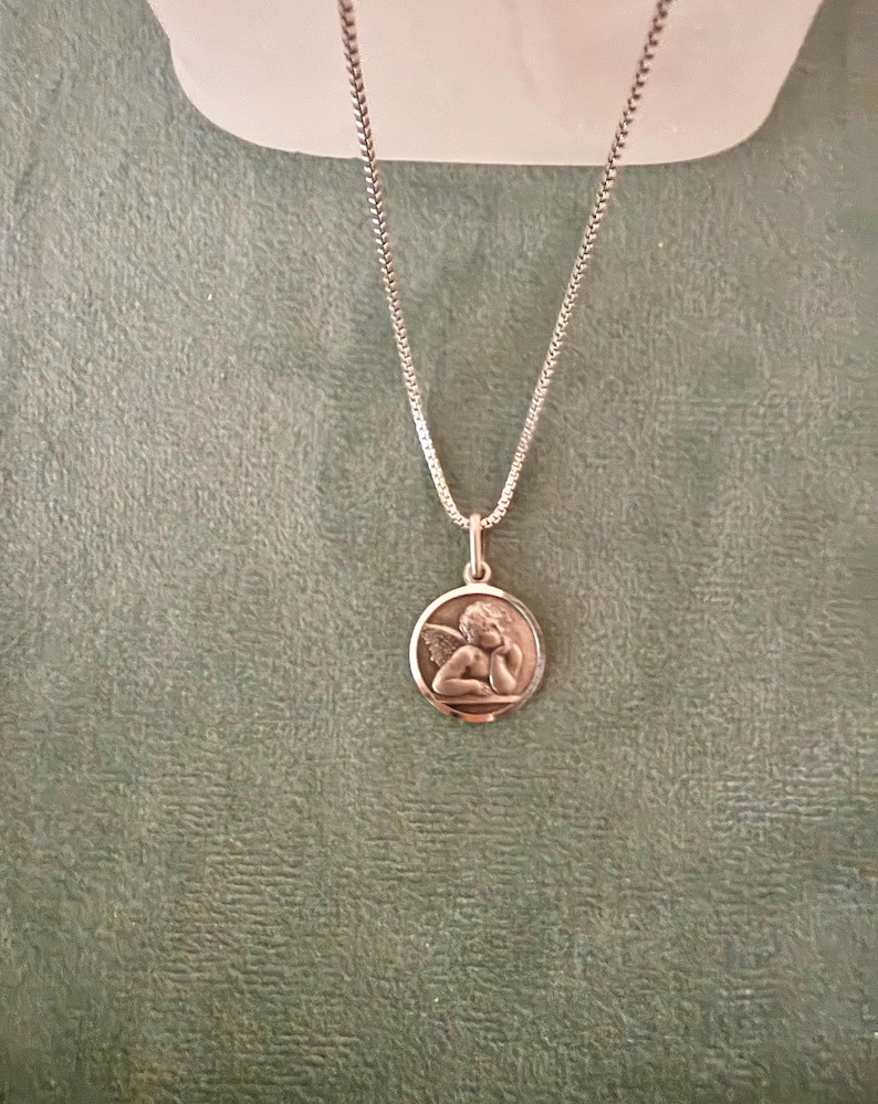 Peut inclure: Collier argent&eacute; avec un pendentif rond repr&eacute;sentant un ch&eacute;rubin en relief. Le ch&eacute;rubin a des ailes et repose sa t&ecirc;te sur sa main. La cha&icirc;ne est de conception fine et tiss&eacute;e. Le pendentif et la cha&icirc;ne sont pos&eacute;s sur un fond vert.