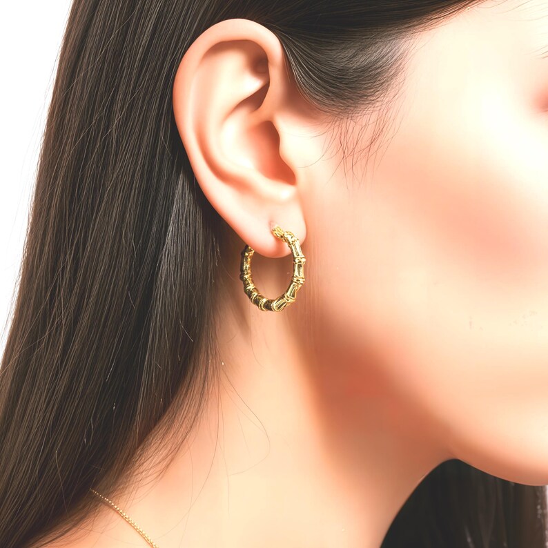 Puede incluir: Un pendiente de aro dorado con un dise&ntilde;o similar al bamb&uacute;. El pendiente se lleva en la oreja de una persona con pelo largo y oscuro. El pendiente tiene una superficie texturizada y forma circular.