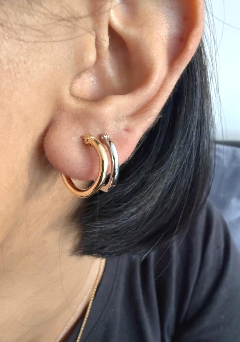 Op de afbeelding: Close-up van twee oorringen. E&eacute;n is goudkleurig en de andere zilverkleurig. De oorbellen worden op het oor gedragen. De gouden oorring zit voor de zilveren oorring. De oorbellen hebben een eenvoudig, klassiek ontwerp.