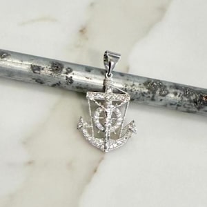 K&ouml;nnte beinhalten: Ein silberfarbener Anh&auml;nger in Form eines Ankers, mit einem Kreuz und einer Figur in der Mitte. Der Anker ist mit kleinen, funkelnden Steinen besetzt. Der Anh&auml;nger h&auml;ngt an einer kleinen Schlaufe.