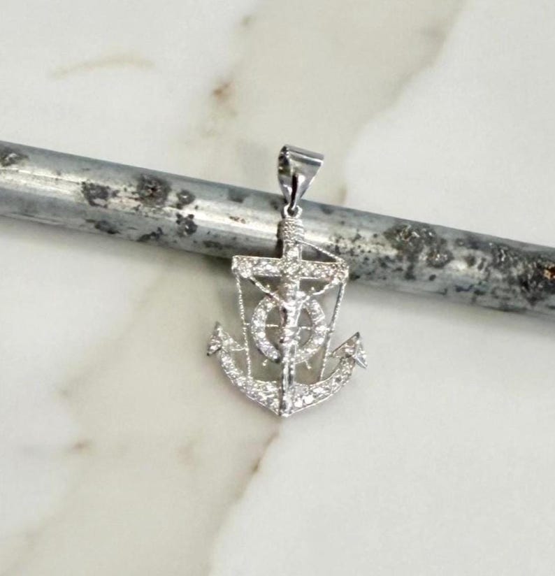 K&ouml;nnte beinhalten: Ein silberfarbener Anh&auml;nger in Form eines Ankers, mit einem Kreuz und einer Figur in der Mitte. Der Anker ist mit kleinen, funkelnden Steinen besetzt. Der Anh&auml;nger h&auml;ngt an einer kleinen Schlaufe.