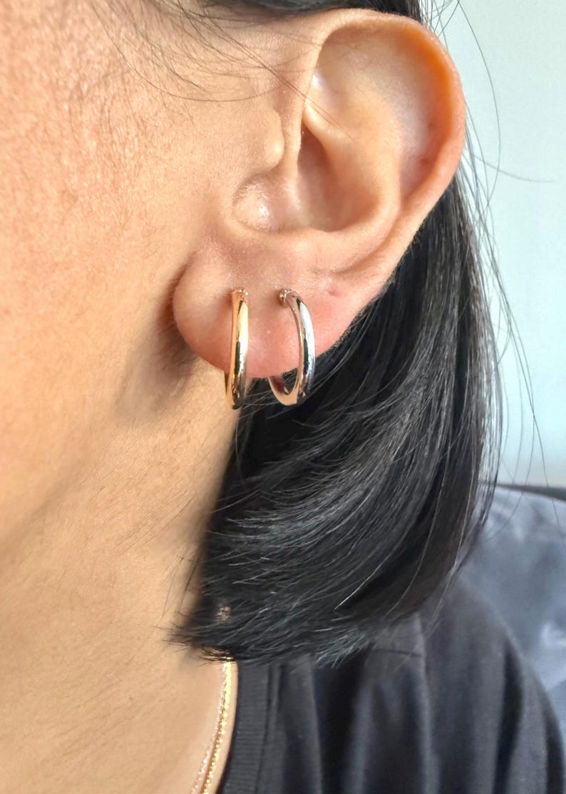 Op de afbeelding: Close-up van een oor met twee oorringen. E&eacute;n oorring is goudkleurig, de andere zilverkleurig. De kleine, cirkelvormige oorringen worden in de oorlellen gedragen.