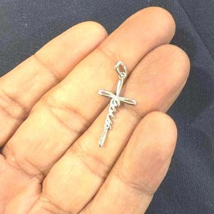 May include: A silver-colored pendant in the shape of a cross, with the word "Jesus" written in a cursive script. The pendant is held in an open hand against a dark background.