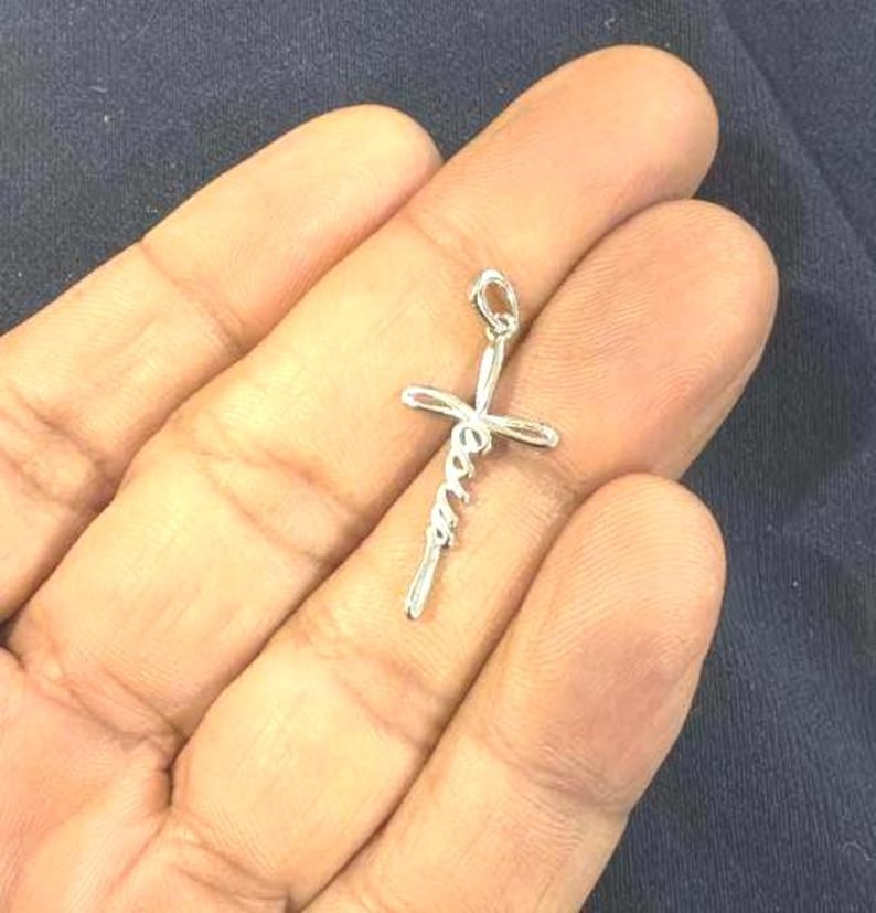 May include: A silver-colored pendant in the shape of a cross, with the word "Jesus" written in a cursive script. The pendant is held in an open hand against a dark background.