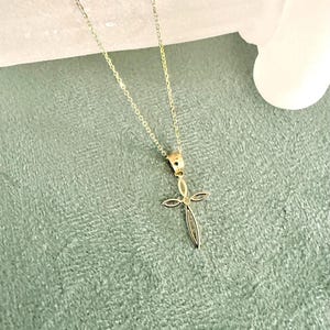 Puede incluir: Un delicado collar con colgante dorado con un dise&ntilde;o de cruz. La cruz tiene una forma delgada y alargada con una peque&ntilde;a gema en el centro. La fina cadena complementa el colgante. El fondo es verde suave y texturizado.