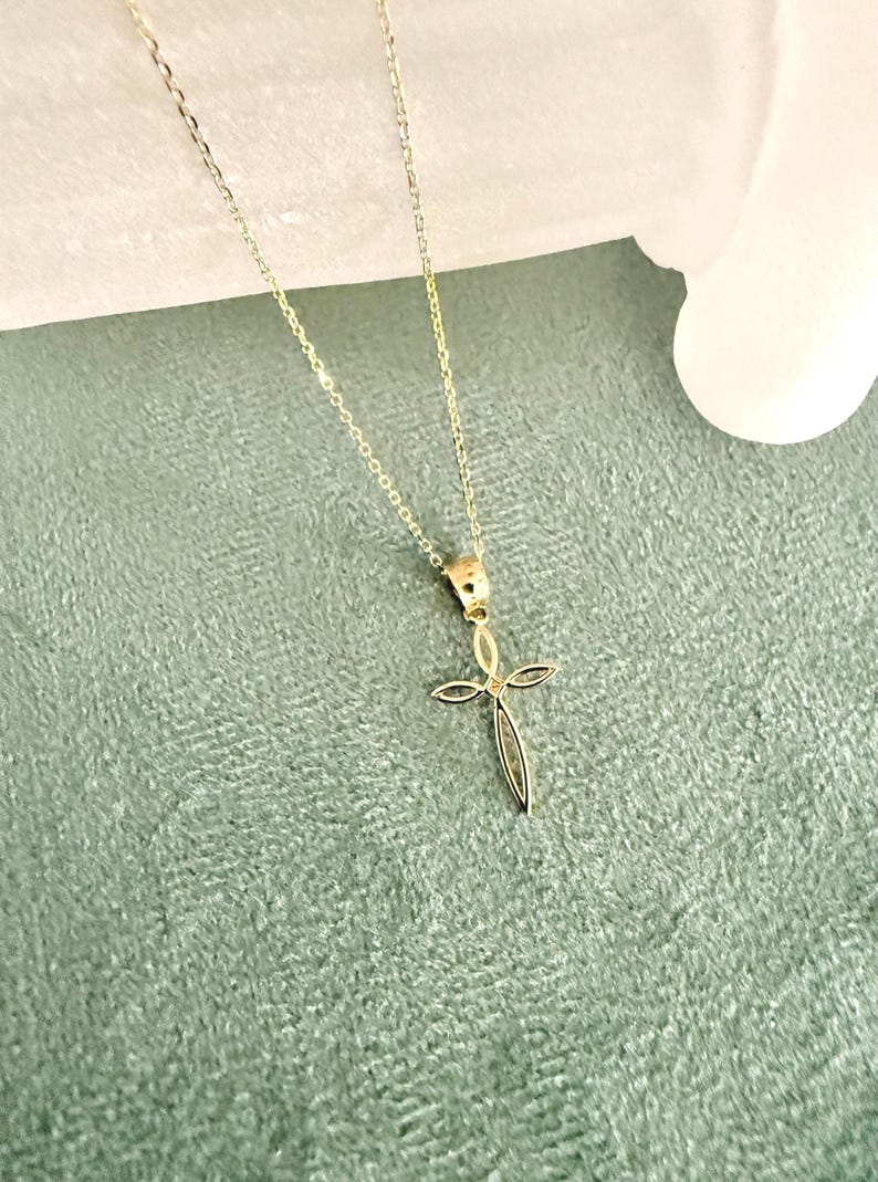 Puede incluir: Un delicado collar con colgante dorado con un dise&ntilde;o de cruz. La cruz tiene una forma delgada y alargada con una peque&ntilde;a gema en el centro. La fina cadena complementa el colgante. El fondo es verde suave y texturizado.
