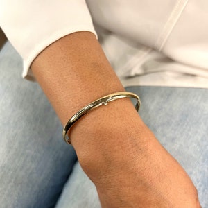 May include: A gold-colored bangle bracelet with a clasp closure. The bracelet is smooth and shiny, reflecting light. The jewelry is worn on a person's wrist, against a background of a white shirt and blue jeans.
