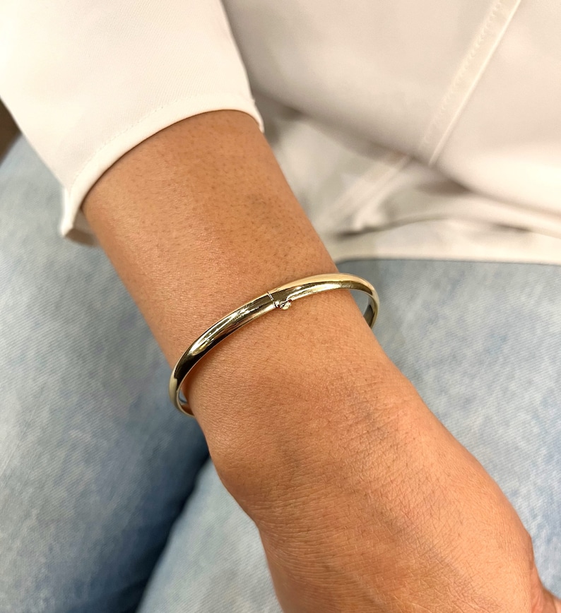 May include: A gold-colored bangle bracelet with a clasp closure. The bracelet is smooth and shiny, reflecting light. The jewelry is worn on a person's wrist, against a background of a white shirt and blue jeans.
