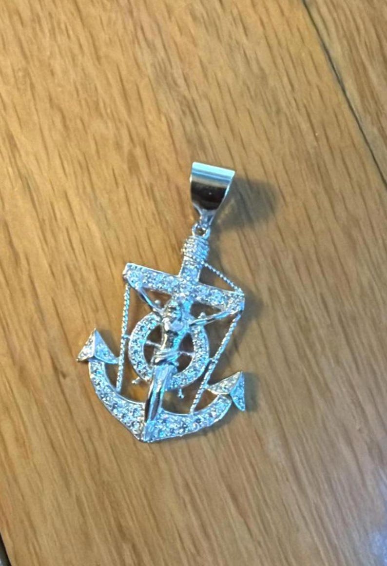 K&ouml;nnte beinhalten: Silberfarbener Anh&auml;nger mit Anker-Design, Kreuz und Figur, verziert mit funkelnden Akzenten. Das Design des Anh&auml;ngers erzeugt einen auff&auml;lligen Kontrast. Ein Schmuckst&uuml;ck mit religi&ouml;sem Thema.