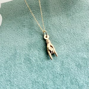 May include: Gold-colored necklace with a pendant shaped like a hand making the 'rock on' gesture. The pendant is suspended from a delicate chain. The background is a solid teal color.