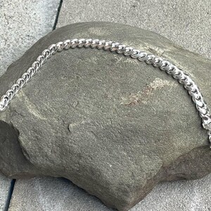 May include: A silver curb chain bracelet with a lobster clasp closure. The bracelet is displayed on a gray stone surface. The chain links are rounded and interlocked, creating a textured appearance. This jewelry item is suitable for everyday wear.