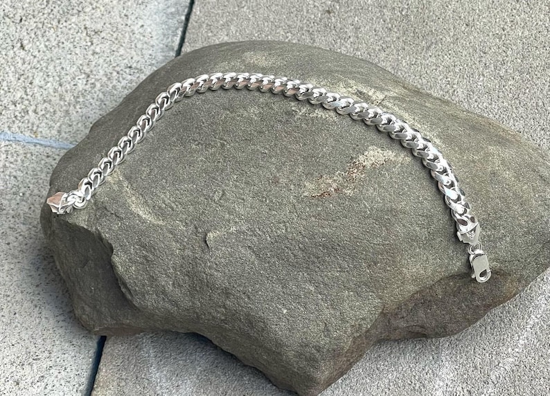 May include: A silver curb chain bracelet with a lobster clasp closure. The bracelet is displayed on a gray stone surface. The chain links are rounded and interlocked, creating a textured appearance. This jewelry item is suitable for everyday wear.
