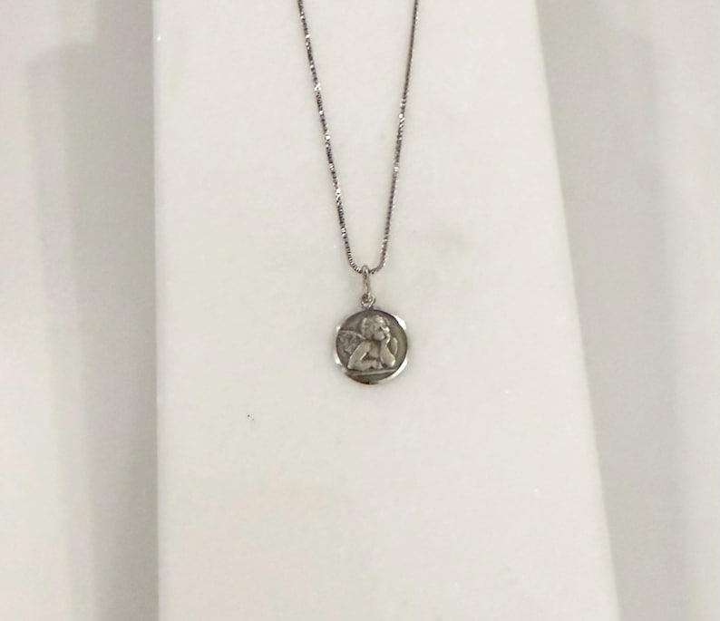 Peut inclure: Un collier argent&eacute; avec un petit pendentif rond. Le pendentif pr&eacute;sente un relief d&eacute;taill&eacute; d'un ange. La cha&icirc;ne est fine et le pendentif est centr&eacute;, cr&eacute;ant un design simple et &eacute;l&eacute;gant.