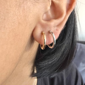 Op de afbeelding: Close-up van een oor met twee oorringen. E&eacute;n oorring is goudkleurig, de andere zilverkleurig. De kleine, ronde oorbellen zitten in de oorlellen. De persoon heeft donker haar. Klassieke sieraden.