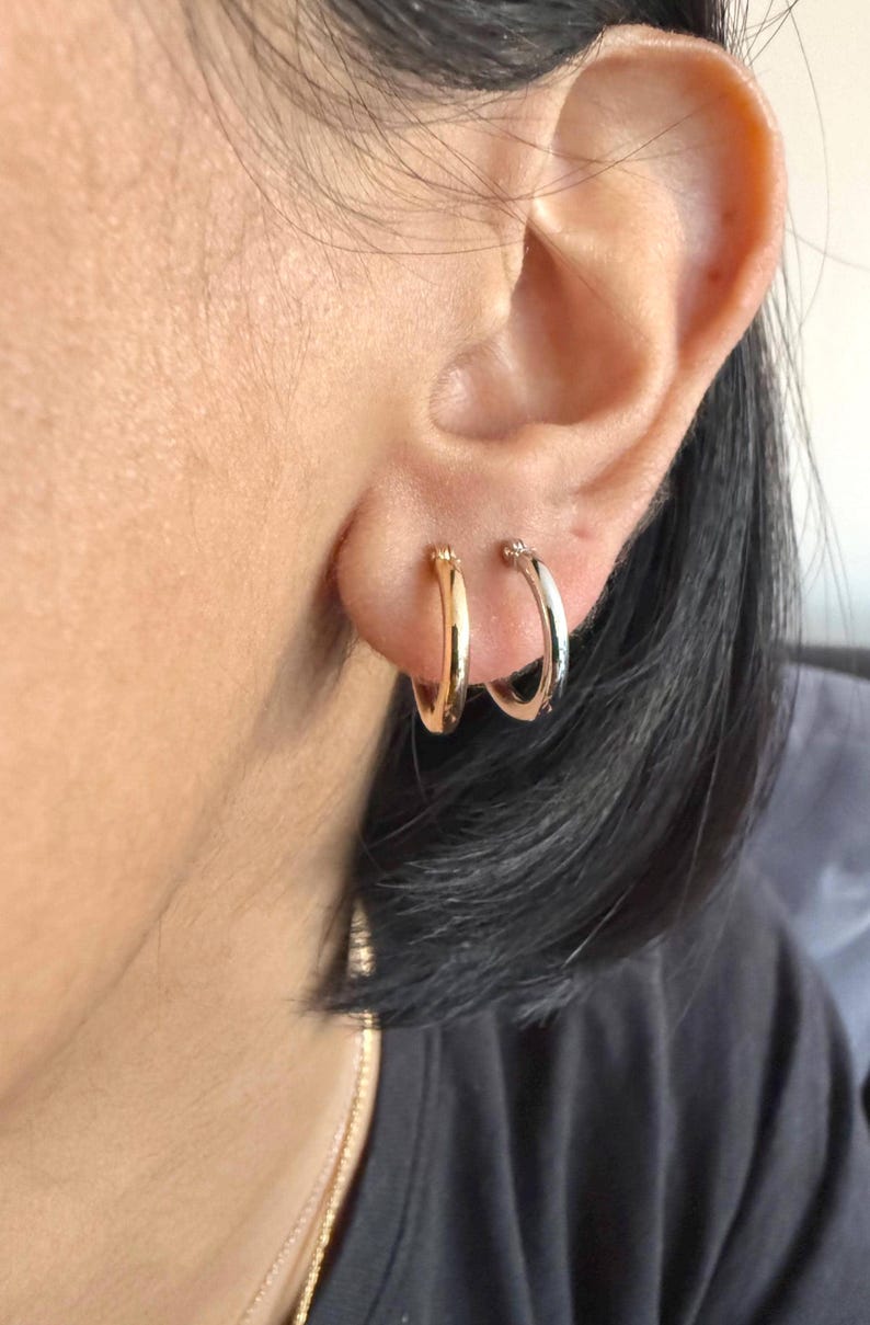 Op de afbeelding: Close-up van een oor met twee oorringen. E&eacute;n oorring is goudkleurig, de andere zilverkleurig. De kleine, ronde oorbellen zitten in de oorlellen. De persoon heeft donker haar. Klassieke sieraden.