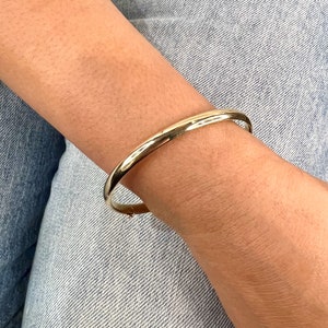 May include: A gold-colored bangle bracelet. The bracelet is a simple, rounded design, and appears to be made of metal. The bracelet is worn on a wrist, with denim jeans in the background.