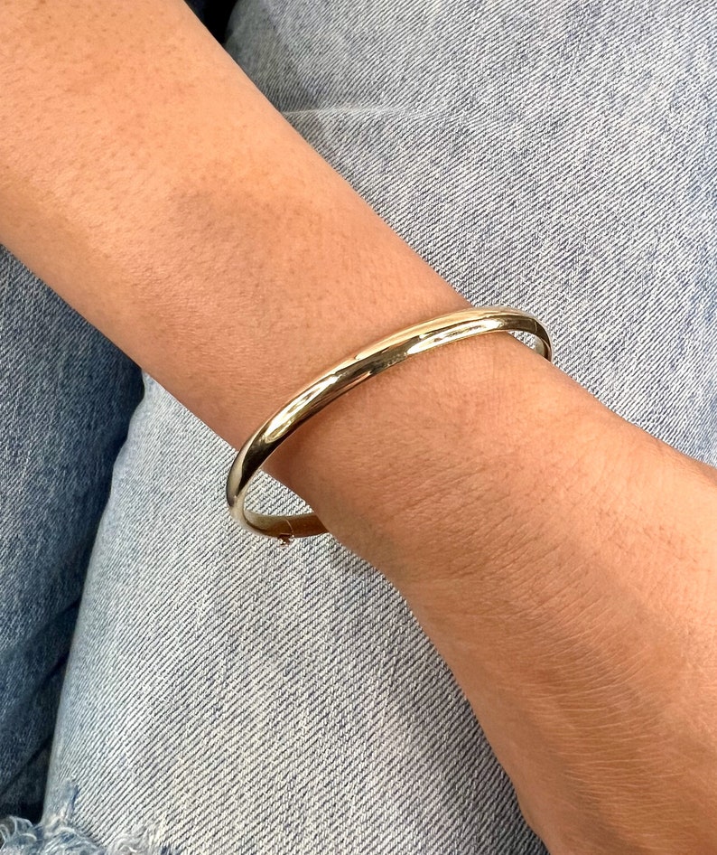 May include: A gold-colored bangle bracelet. The bracelet is a simple, rounded design, and appears to be made of metal. The bracelet is worn on a wrist, with denim jeans in the background.