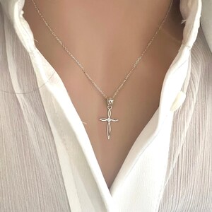 Puede incluir: Un collar con colgante de cruz plateada. La cruz tiene un dise&ntilde;o calado con un peque&ntilde;o acento redondo en el centro. El collar se lleva sobre una camisa blanca.